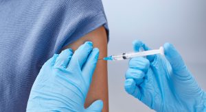 Healthcare worker injects a vaccine into a patient’s upper arm, both wearing blue gloves (close-up).