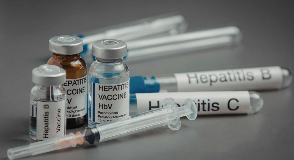 Close-up of vaccine vials labeled Hepatitis B and other vaccines with syringes on a gray background.