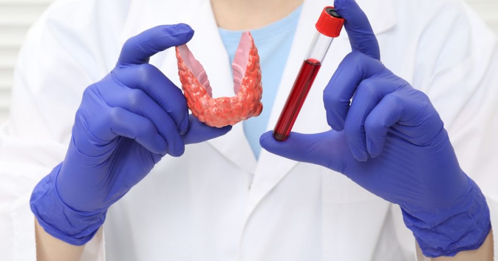 Scientist wearing blue gloves holds a pink dental jaw model and a blood sample in a vial against a lab coat background, illustrating dental research.
