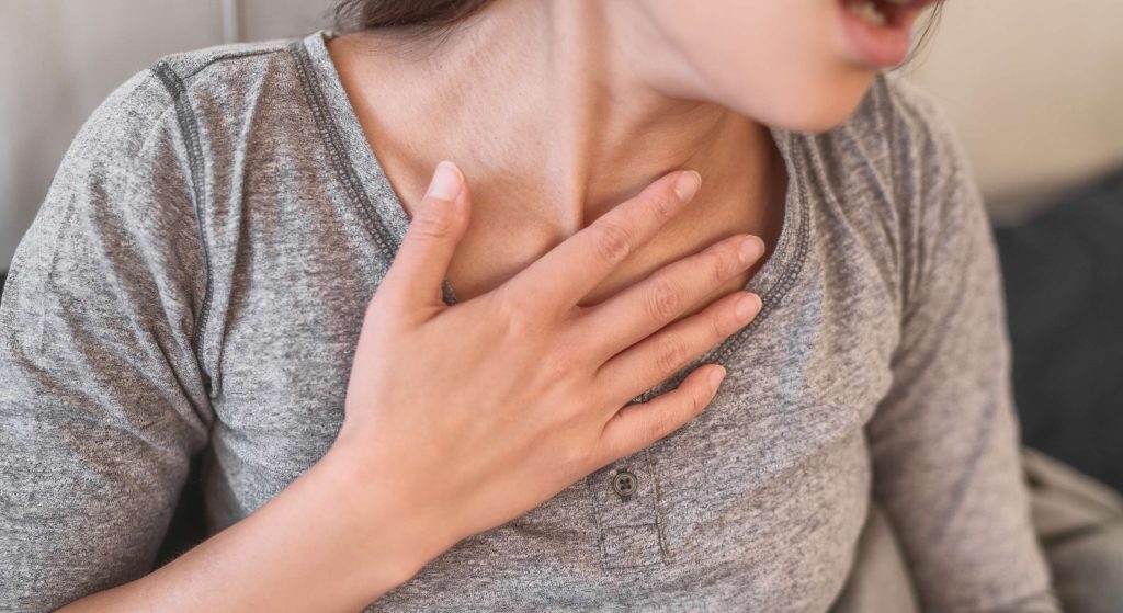 Person in a gray shirt touching their neck with one hand, suggesting throat discomfort or pain.