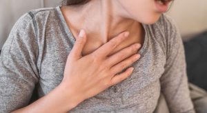Person in a gray shirt touching their neck with one hand, suggesting throat discomfort or pain.